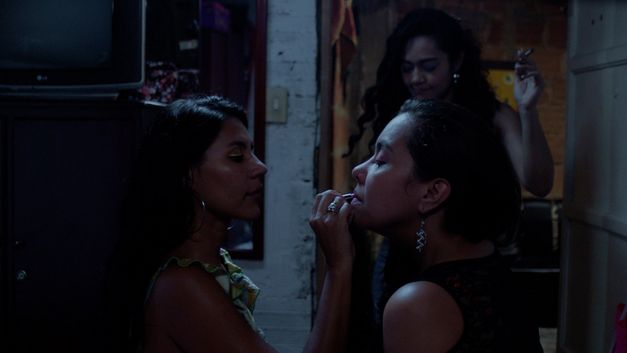 Film still from "La piel en primavera" by Yennifer Uribe Alzate. It shows three women. A woman on the left is applying make-up to the lips of the woman on the right. The third woman comes through the door in the background with a cigarette in her hand. 