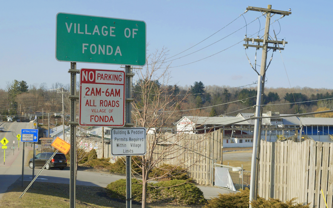 Film still from HENRY FONDA FOR PRESIDENT: It shows the entrance sign to the village of Fonda. A road and houses can be seen in the background. 