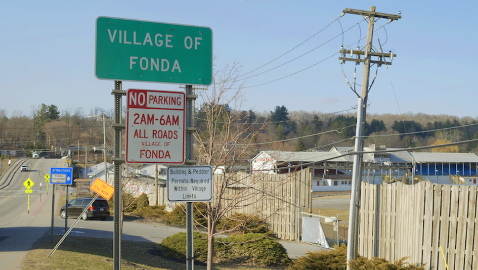 Film still from HENRY FONDA FOR PRESIDENT: It shows the entrance sign to the village of Fonda. A road and houses can be seen in the background. 