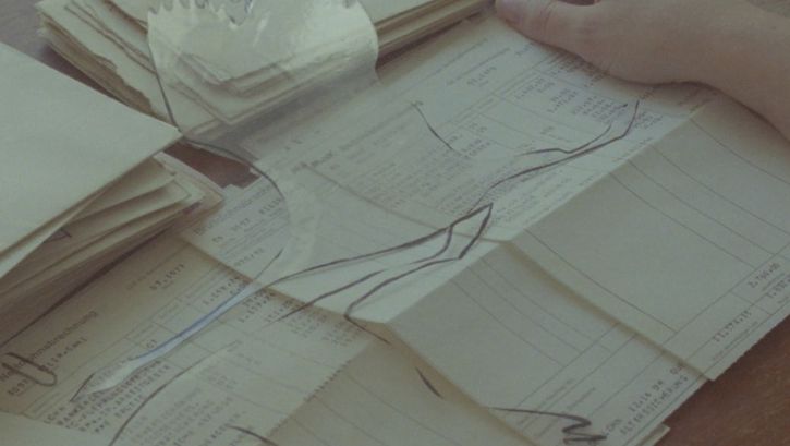 Filmstill aus dem Film „Zwischenwelt“ von Cana Bilir-Meier. Die Hände einer Person berühren einen Stapel Lohnabrechnungen. Über dem Papierstapel zwei, in transparentes Material ausgeschnittene fliegende Vögel. 