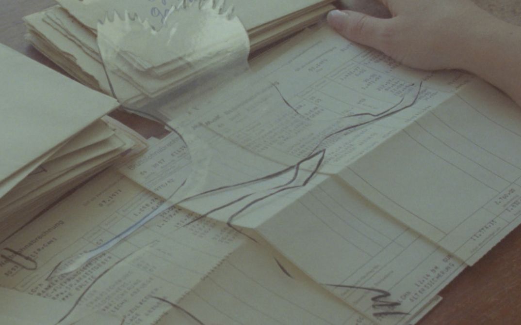 Filmstill aus dem Film „Zwischenwelt“ von Cana Bilir-Meier. Die Hände einer Person berühren einen Stapel Lohnabrechnungen. Über dem Papierstapel zwei, in transparentes Material ausgeschnittene fliegende Vögel.