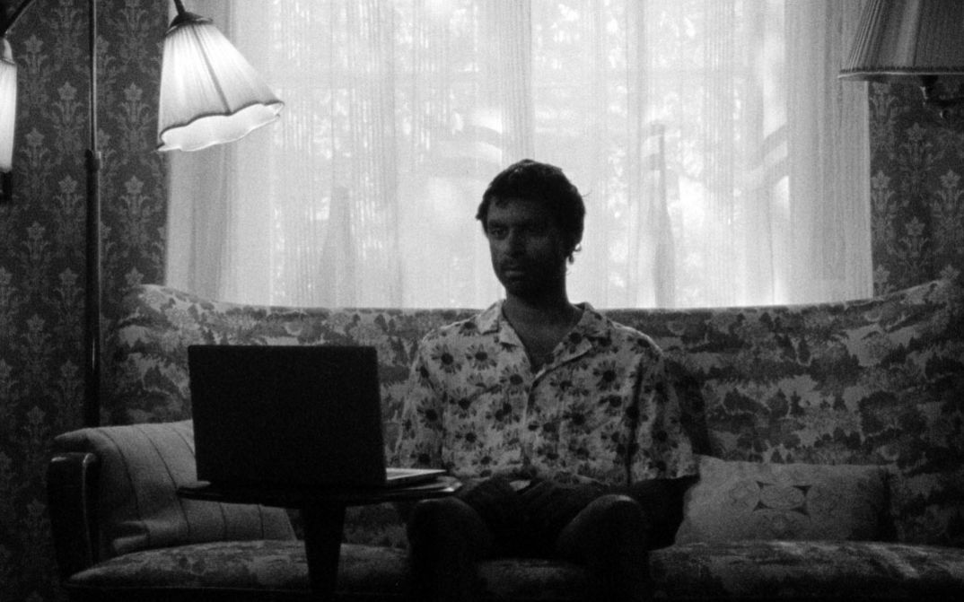 Person sitzt auf einem Sofa, vor sich ein aufgeklappter Laptop, im Hintergrund ein Fenster mit Vorhängen.