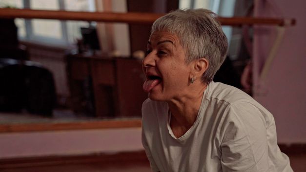 Film still from NAGOTA by Sabina Bakaeva. The image shows a woman with short gray hair sticking out her tongue. 