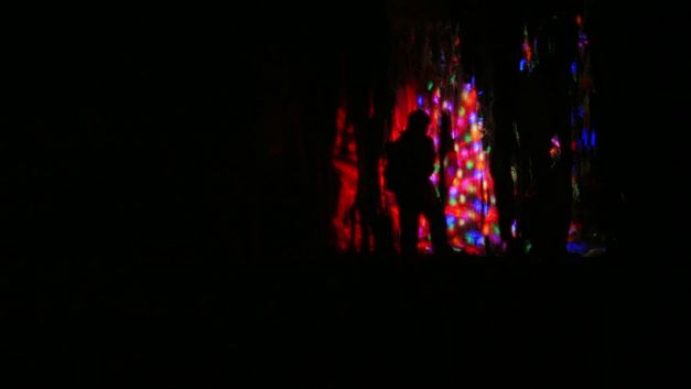 Film still from Chonchanok Thanatteepwong and Pobwarat Maprasob’s film “Mountain Roars”. A dark silhouette standing out in the shadows of a cave, bathed in a kaleidoscope of colored lights.