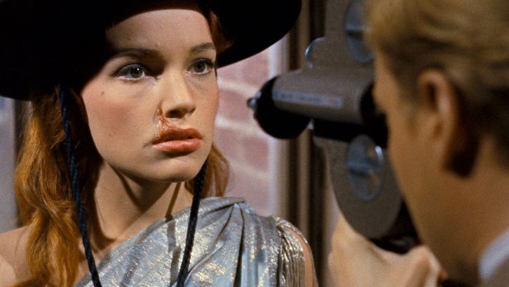 Filmstill aus PEEPING TOM: Eine junge Frau mit langen Haaren, einem schwarzen Hut und silbrig glänzenden Kleid wird von einem direkt vor ihr stehendem Mann gefilmt. Sie hat eine Narbe an der Oberlippe.