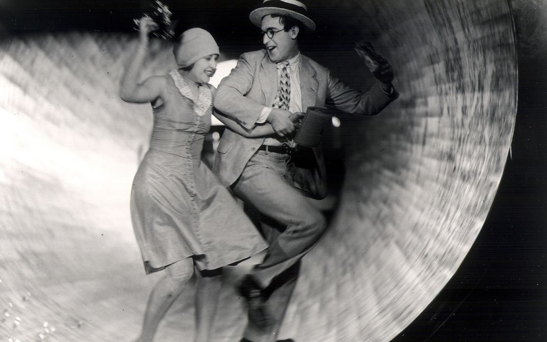 Film still from SPEEDY from 1928: A man and a woman are standing inside a tube-like structure at an amusement park, smiling at each other.