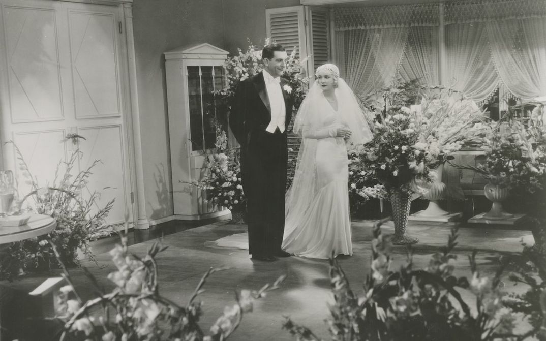 Filmstill aus DESIGN FOR LIVING: Ein Hochzeitspaar steht in einem Zimmer voller Blumen.
