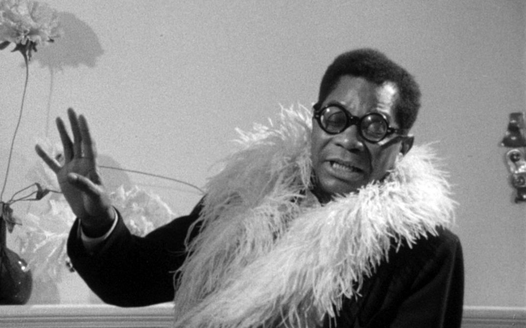Film still from PORTRAIT OF JASON: A man with glasses and a feather boa in conversation.