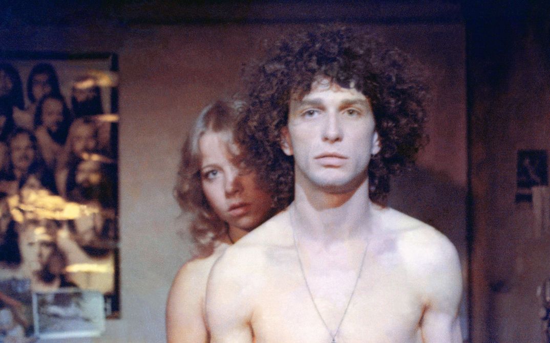 Filmstill aus RUMMEL: Ein Mann und eine Frau, oberkörperfrei, er mit einer Kreuzkette um den Hals, blicken recht ausdruckslos in die Kamera. An der Wand sind Poster und Fotos.