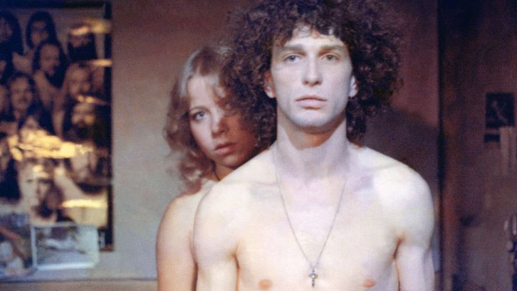 Filmstill aus RUMMEL: Ein Mann und eine Frau, oberkörperfrei, er mit einer Kreuzkette um den Hals, blicken recht ausdruckslos in die Kamera. An der Wand sind Poster und Fotos.