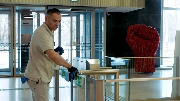 Film still from "For the Many – The Vienna Chamber of Labour" by Constantin Wulff: In the entrance hall of an office building, a man is cleaning the gate. To the right, there is a sculpture: A big, red fist.