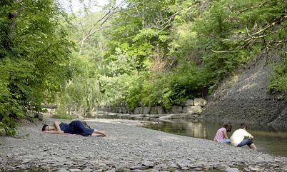 Filmstill from „Concrete Valley" by Antoine Bourges. in a bucolic landscape two children are playing on the bank of a river and a woman is sleeping nearby.