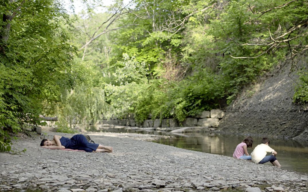 Filmstill aus „Concrete Valley" von Antoine Bourges. In einer bukolischen Landschaft spielen zwei Kinder am Ufer eines Flusses und eine Frau schläft in der Nähe.
