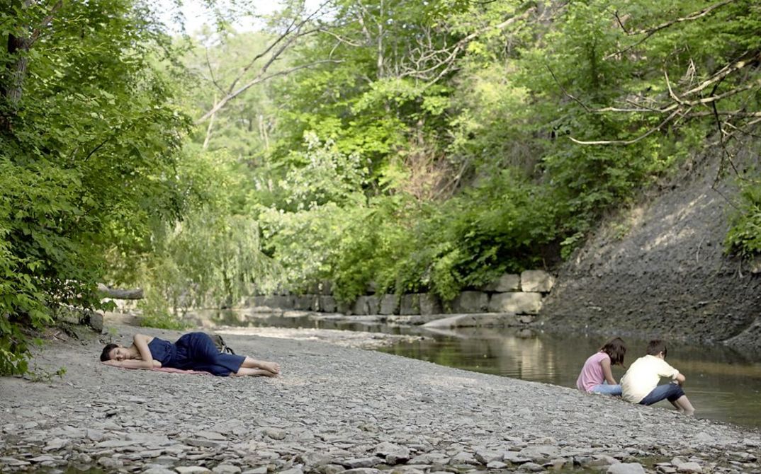 Filmstill from „Concrete Valley" by Antoine Bourges. in a bucolic landscape two children are playing on the bank of a river and a woman is sleeping nearby.