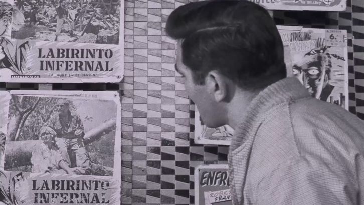 Film still from BELARMINO: A dark-haired man looking away from the camera at a wall with posters. The poster have the writing  “Labirinto Infernal” on them.