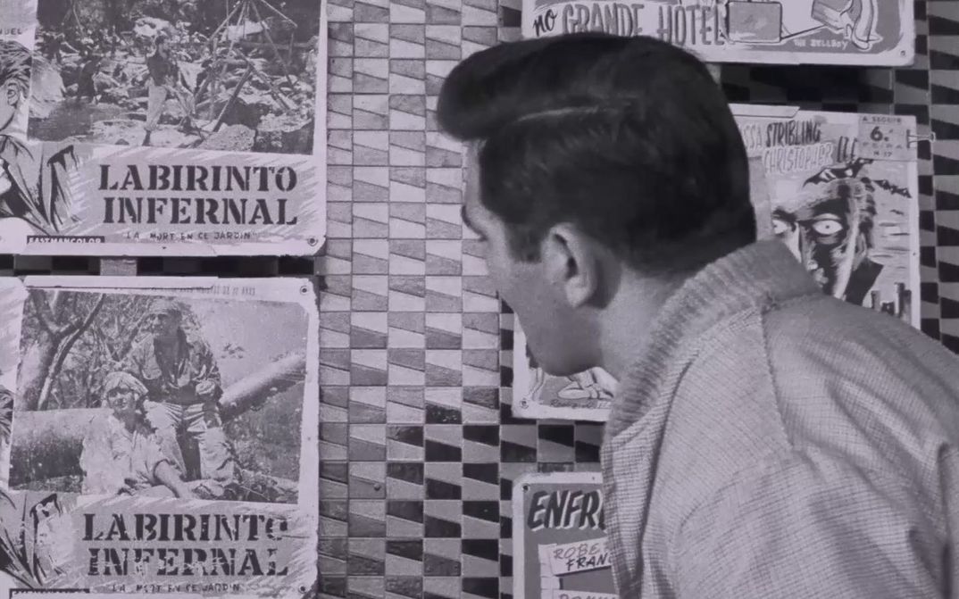 Film still from BELARMINO: A dark-haired man looking away from the camera at a wall with posters. The poster have the writing  “Labirinto Infernal” on them.