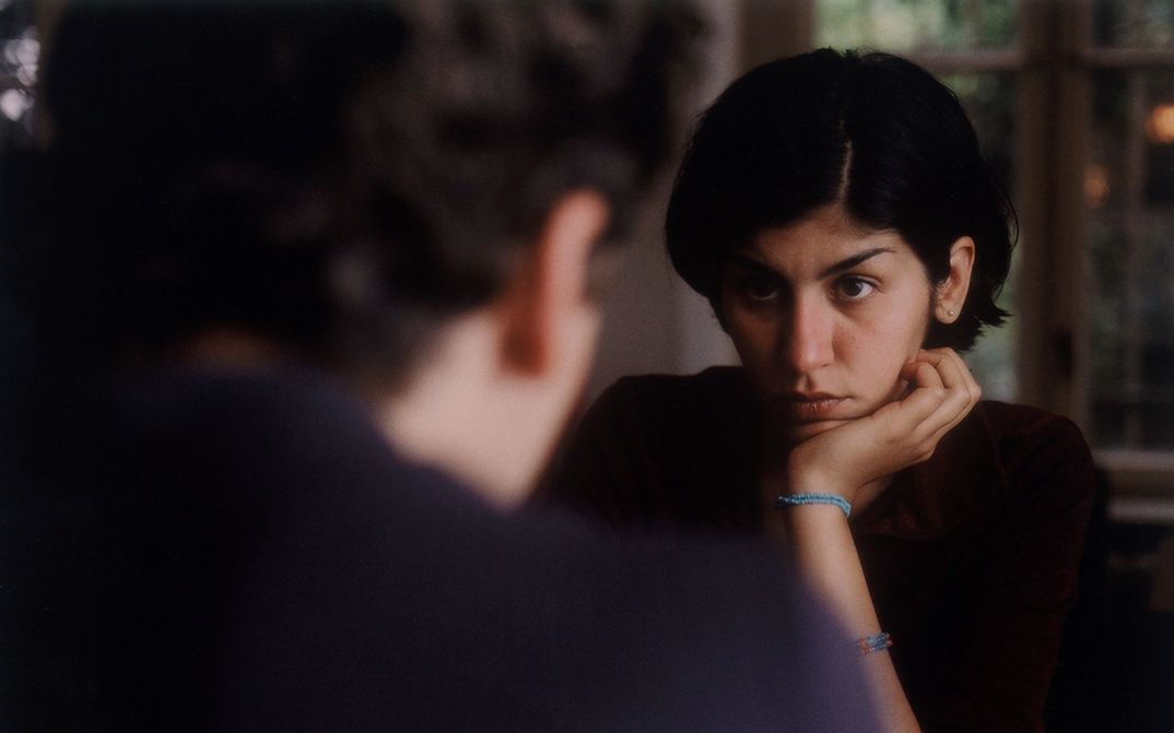 Filmstill aus DER SCHÖNE TAG: Zwei Personen sitzen sich gegenüber. Die eine Person ist nur von hinten zu sehen und die andere Person von vorne, sie hat ihr Kinn auf die Hand gestützt. Die Personen schauen sich an.