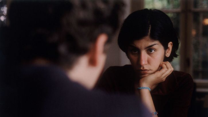 Film still from THE BEAUTIFUL DAY: Two people are sitting opposite each other. One person can only be seen from behind and the other from the front, leaning their chin on their hand. The people look at each other.