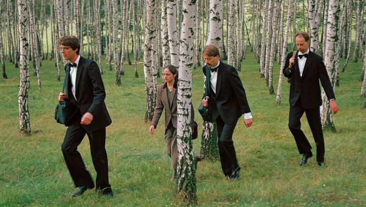 Filmstill aus DIE DREI GERECHTEN KAMMACHER: 3 Männer in Anzug und Fliege und eine Frau in braunem Anzug laufen in einer Reihe durch einen Birkenhain.
