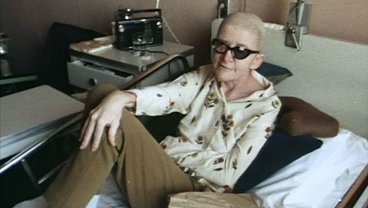 Film still from DYING: A person is sitting in a hospital bed.