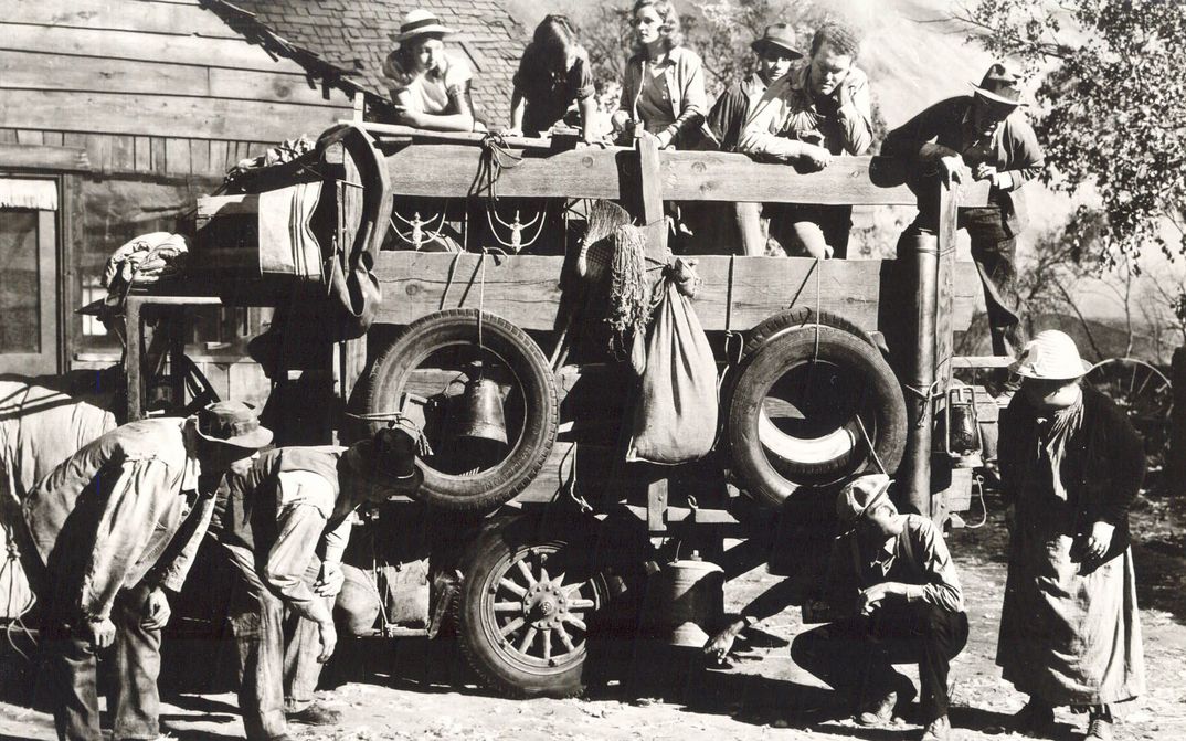 Filmstill aus THE GRAPES OF WRATH: Eine Gruppe von Menschen steht auf und vor einem beladenen Planwagen und begutachten die Reifen.