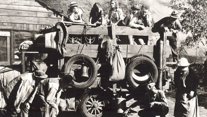 Filmstill aus THE GRAPES OF WRATH: Eine Gruppe von Menschen steht auf und vor einem beladenen Planwagen und begutachten die Reifen.
