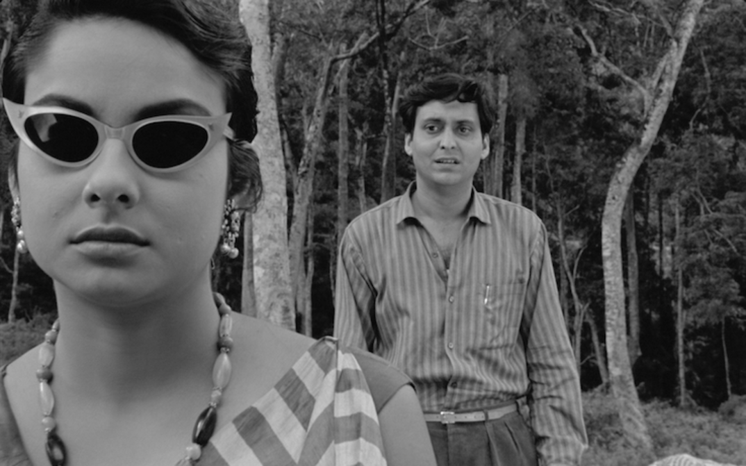 Filmstill aus KAPURUSH: Ein Mann und eine Frauen stehen zwischen Bäumen. Sie trägt eine Sonnenbrille, er steht hinter ihr und schaut sie an.