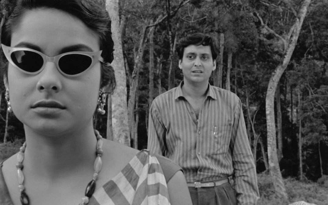 Film still from KAPURUSH: A man and a woman are standing between trees. She is wearing sunglasses, he is standing behind her and looking at her.