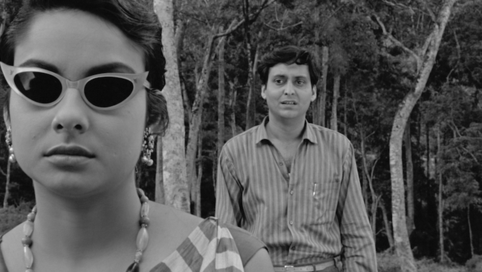 Filmstill aus KAPURUSH: Ein Mann und eine Frauen stehen zwischen Bäumen. Sie trägt eine Sonnenbrille, er steht hinter ihr und schaut sie an.