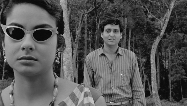 Film still from KAPURUSH: A man and a woman are standing between trees. She is wearing sunglasses, he is standing behind her and looking at her.