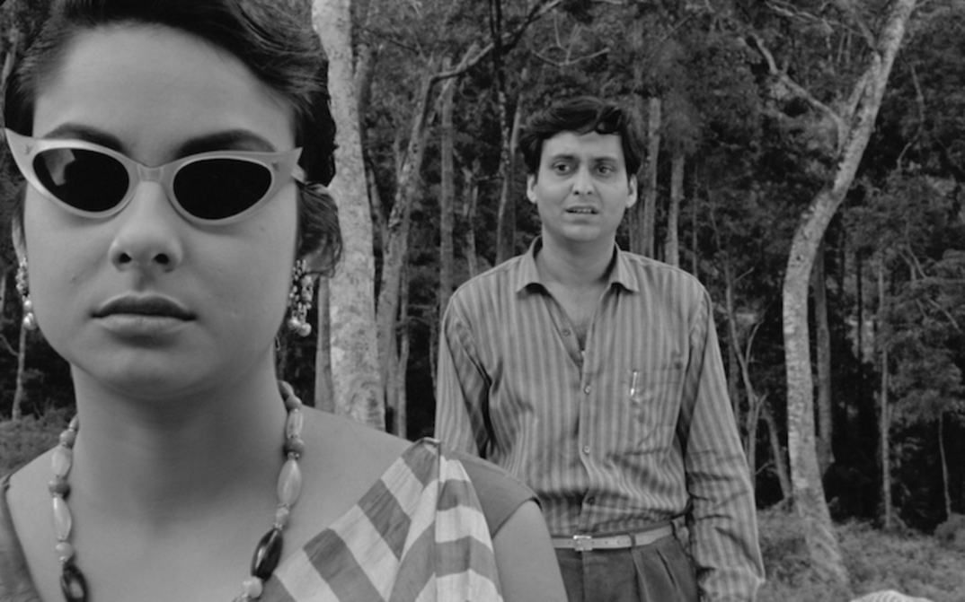 Film still from KAPURUSH: A man and a woman are standing between trees. She is wearing sunglasses, he is standing behind her and looking at her.