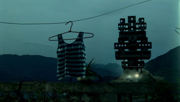 Filmstill aus STILL LIFE: Ein gestreiftes Tanktop hängt am Bügel. Daneben eine Silhouette eines geometrischen Gebildes. Im Hintergrund Berge. Es ist neblig und fast dunkel.