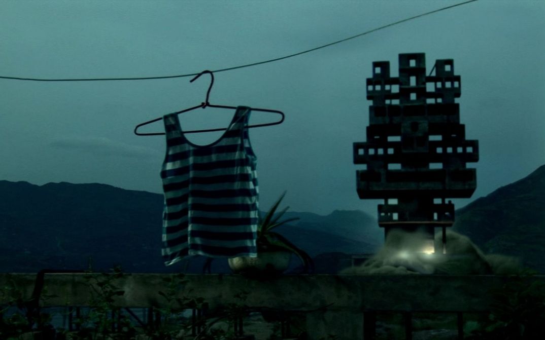 Filmstill from STILL LIFE: A striped tank top hangs on the hanger. Next to it is a silhouette of a geometric shape. Mountains in the background. It is foggy and almost dark.