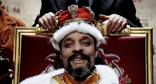 Filmstill aus „West Indies" von Med Hondo. Nahaufnahme eines Mannes mit Ziegenbart auf einem Thron. Er trägt eine rote Krone und lächelt. 