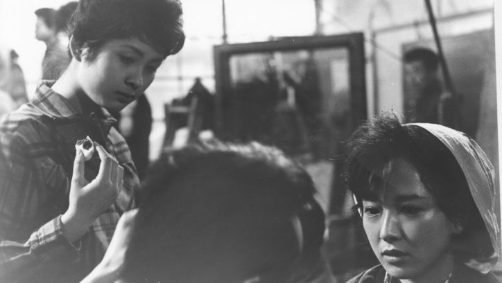 Filmstill aus A FULL LIFE: Man sieht drei Personen im Gespräch. Im Hintergrund sind weitere Menschen zu sehen.