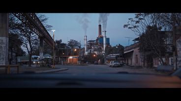 A street at dawn with a truck driving into the frame from the left, in the background there is a factory building with smoking chimneys.