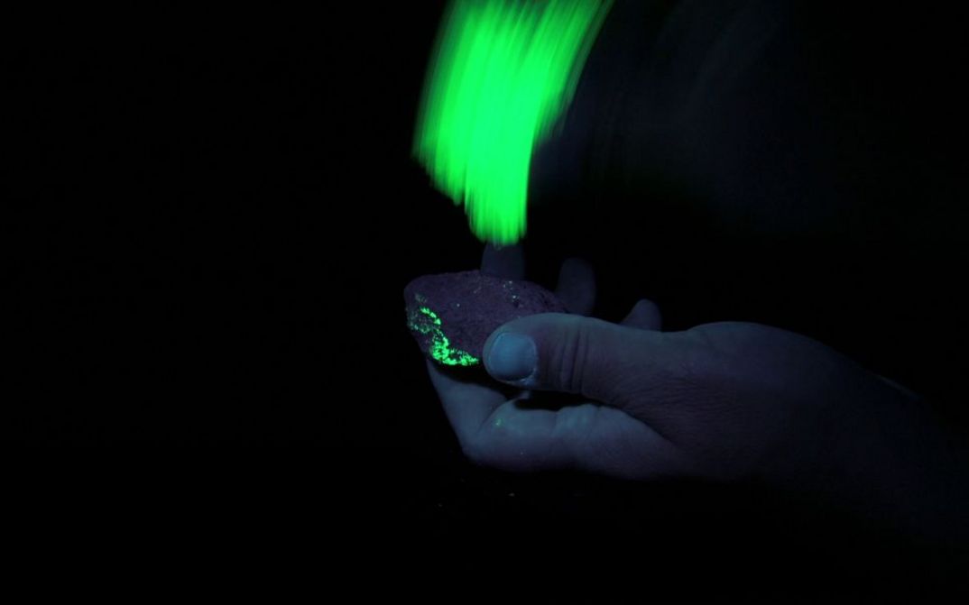 Filmstill aus dem Film "Sonne Unter Tage" von Mareike Bernien und Alex Gerbaulet. Eine Hand hält einen fluoriszierenden Stein der von Schwarzlich beleuchtet wird.