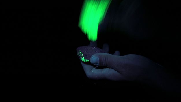 Still from the film "Sun Under Ground" by Mareike Bernien and Alex Gerbaulet. A hand holds a fluorescent stone illuminated by black light.