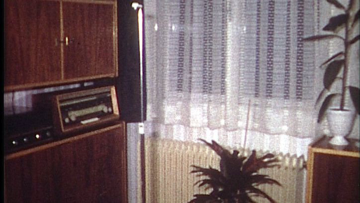 Filmstill aus EINE MILLION KREDIT IST NORMAL, SAGT MEIN GROSSVATER: Man sieht eine Ecke eines Wohnzimmers mit Schrankwand und einem Radio, einen Heizkörper unter dem Fenster und einem weißen Spitzenvorhang und zwei Topfpflanzen.