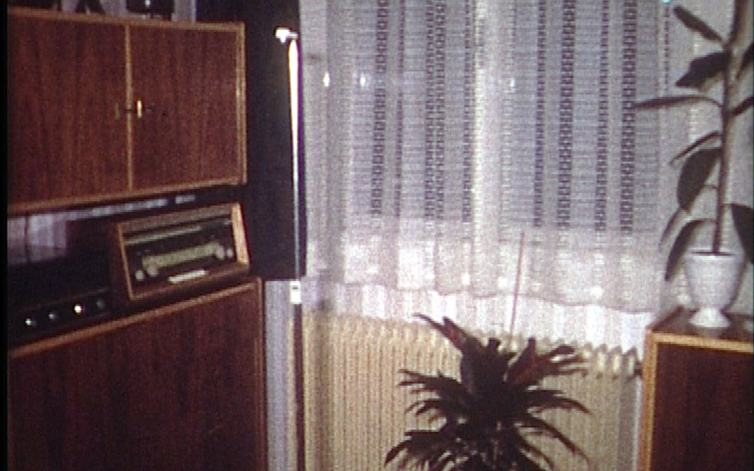 Film still from EINE MILLION KREDIT IST NORMAL, SAGT MEIN GROSSVATER: You can see a corner of a living room with a wall unit and a radio, a radiator under the window, a white lace curtain, and two potted plants.