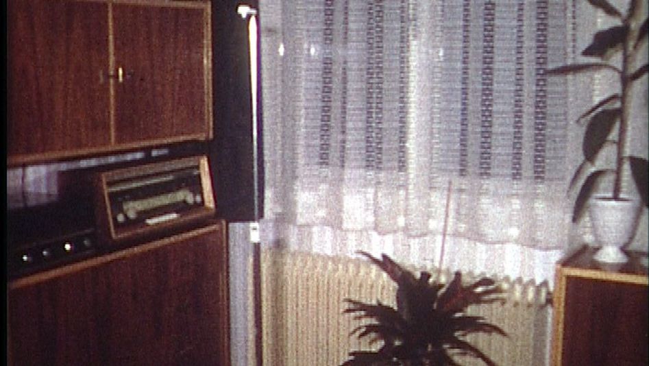 Film still from EINE MILLION KREDIT IST NORMAL, SAGT MEIN GROSSVATER: You can see a corner of a living room with a wall unit and a radio, a radiator under the window, a white lace curtain, and two potted plants.