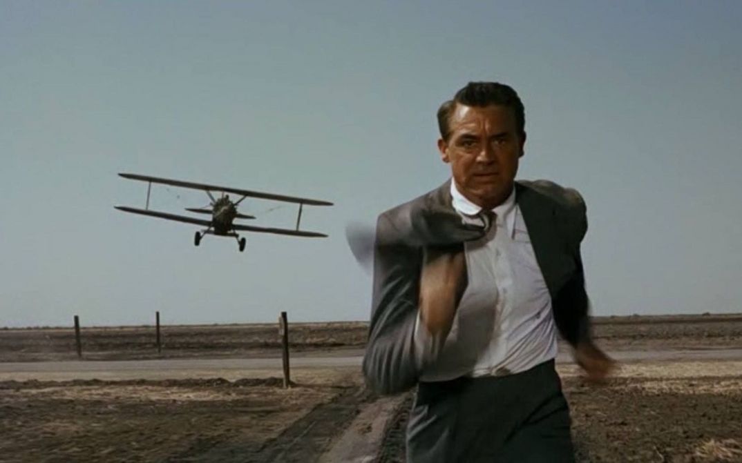Filmstill aus NORTH BY NORTHWEST: Ein Mann im Anzug rennt über ein Feld, er wird von einem kleinen Flugzeug verfolgt.