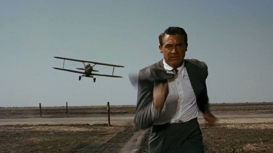 Filmstill aus NORTH BY NORTHWEST: Ein Mann im Anzug rennt über ein Feld, er wird von einem kleinen Flugzeug verfolgt.