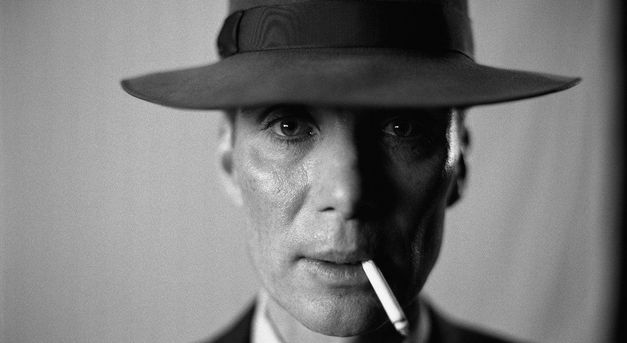 Film still from OPPENHEIMER: Close-up of the actor Cillian Murphy as Oppenheimer. He is wearing a hat, has a cigarette in the corner of his mouth and is looking head-on into the camera.