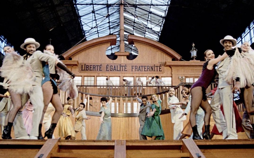 Filmstill aus „West Indies" von Med Hondo. Eine Gruppe von Tänzern und Tänzerinnen stehen auf der Bühne. Die Kulisse besteht aus einem Sklavenschiff, auf dem die Aufschrift „Liberté Égalité, Fraternité“ zu sehen ist.