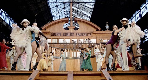 Still from the film "West Indies" by Med Hondo. We see a group of men and women dancers facing the camera while standing on a replica slave ship bearing the words "Liberté Égalité, Fraternité"