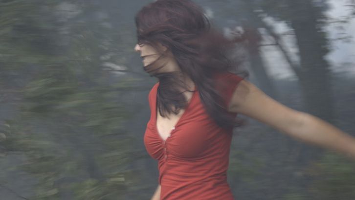 Film still from ÇA BRÛLE: A young woman with long brown hair and a red top is standing in front of a patch of woods, which you can only see out of focus. Her face is half hidden by her hair.