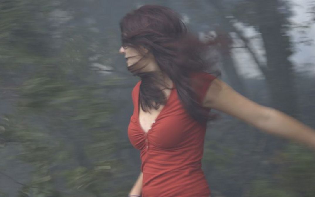 Filmstill aus ÇA BRÛLE: Eine junge Frau mit langen braunen Haaren und einem roten Oberteil steht vor einem Waldstück, das man nur unscharf sieht. Ihr Gesicht ist halb durch ihre Haare verdeckt.