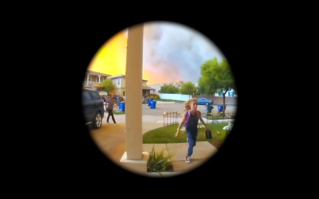 Filmstill from Graeme Arnfield’s „Home Invasion“. A circular view on a front yard. A pillar in the middle, on its right a child, on its left another person and a car. In the background a lit sky and a lot of smoke.