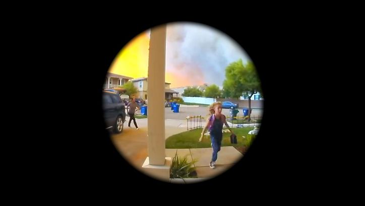 Filmstill aus dem Film „Home Invasion“ von Graeme Arnfield. Ein kreisrunder Blick auf einen Vorgarten. In der Mitte eine Säule, rechts davon ein Kind, links ein Auto und eine weitere Person. Im Hintergund Rauch und helles Leuchten am Himmel.